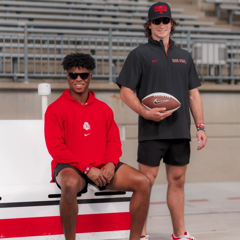 Two men in Ohio State athletic apparel, one sitting wearing a red hoodie and sunglasses, the other standing with a football, both posing in an empty stadium.