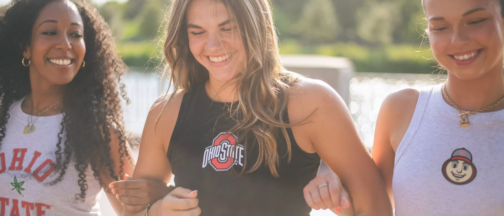 Three women walking together outdoors, wearing Ohio State apparel, smiling and linking arms.